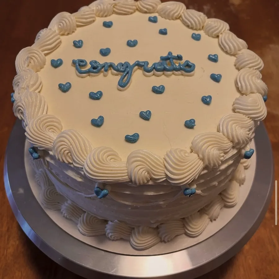 A white cake with blue piped hearts and the word Congrats piped across the top