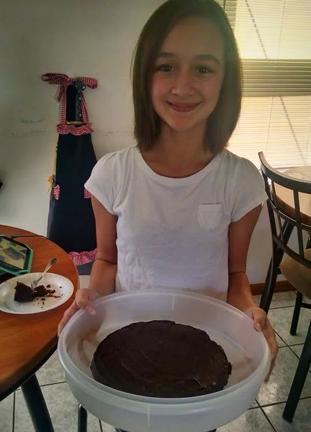A young Ava holding up a round chocolate-frosted cake in a plastic cake carrier