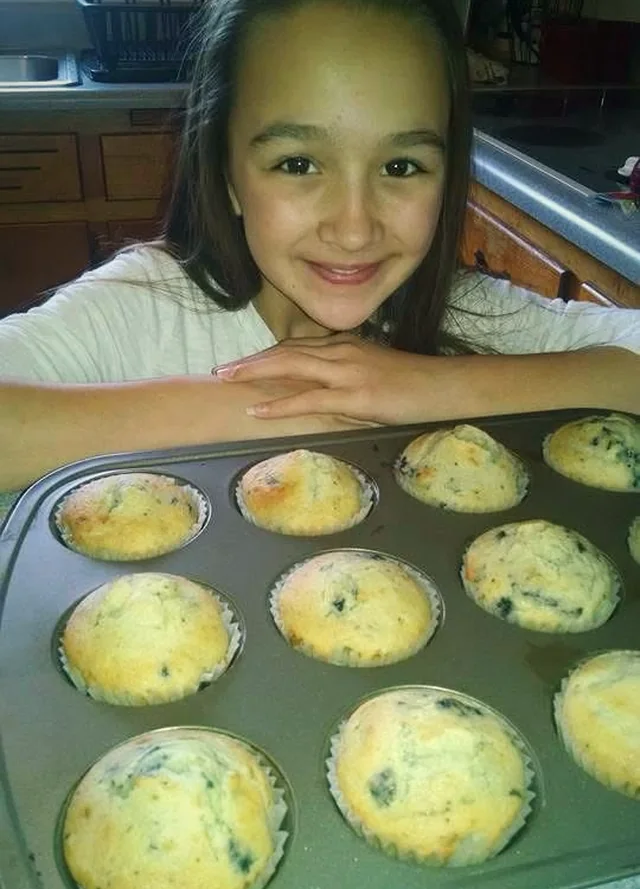 A young Ava resting her chin on her hands behind a tray of freshly baked blueberry muffins